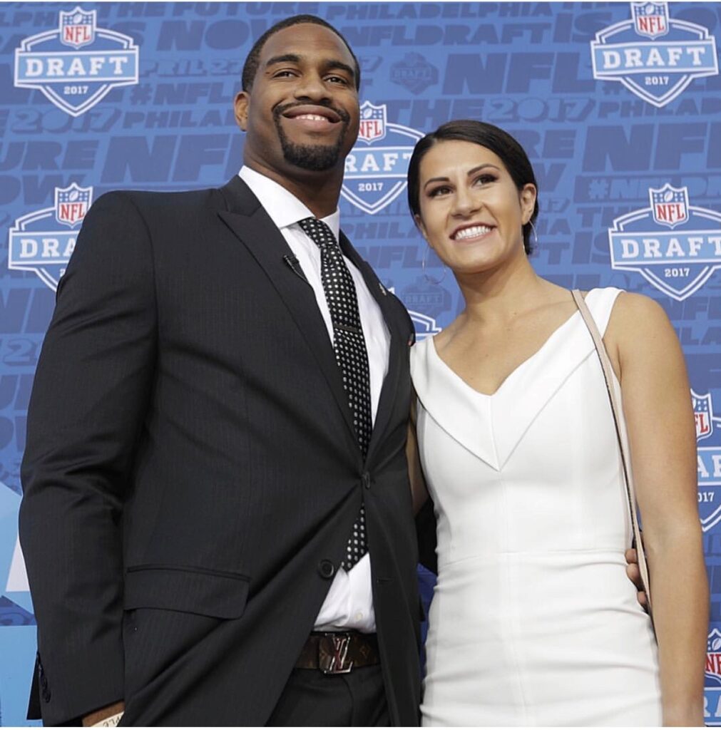 
In this image, a man and a woman are posing together at an event. The man is wearing a black suit with a white dress shirt and a dotted tie. He has a neat beard and a broad smile. The woman is dressed in a white sleeveless dress and has her dark hair pulled back. She is also smiling warmly. They are standing against a blue backdrop that features "NFL Draft 2017" logos and text. The setting appears to be a formal or celebratory event, likely related to the NFL Draft.